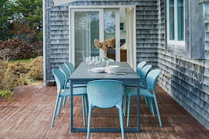 An outdoor dining area with a table set for six, surrounded by six blue chairs, and a sliding glass door leading to a room. - Sun-filled escape with hot tub, deck, bikes, grill, W/D, & Central AC (Truro)
