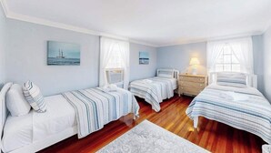 Une chambre avec trois lits, une table de chevet, une lampe et un tableau représentant un voilier.