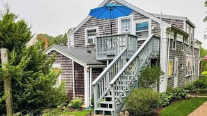 Une maison à deux étages avec un escalier menant à un balcon, le tout entouré de verdure.