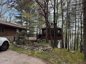 A cabin with a stone foundation, a wooden deck, and a view of a lake surrounded by trees. - Stack's Back Bay Retreat (Minocqua)