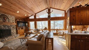 A cabin interior with a stone fireplace, wooden ceiling, a television, a sofa, a dining table, and a kitchen area. - Stack's Back Bay Retreat (Minocqua)