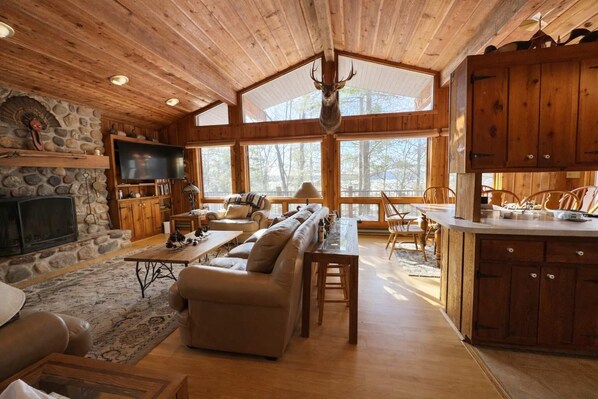 A cabin interior with a stone fireplace, wooden ceiling, a television, a sofa, a dining table, and a kitchen area. - Stack's Back Bay Retreat (Minocqua)