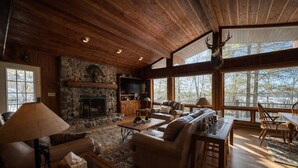 A cozy cabin interior with a stone fireplace, leather sofas, a dining area, and large windows. - Stack's Back Bay Retreat (Minocqua)