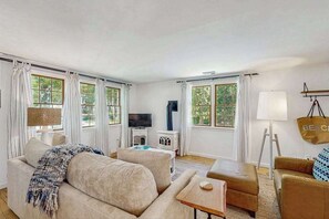 Un salon comprenant un canapé, un pouf, une télévision, une lampe et des fenêtres avec des rideaux.