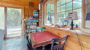 A wooden cabin interior with a dining table, chairs, a bookshelf, and a window with a thermometer. - Waterfront gem with deck, bonus den & 2 kayaks - near dining & beach (South Yarmouth)