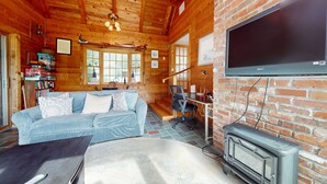 A cozy cabin interior with a sofa, a TV, a fireplace, and a bookshelf. - Waterfront gem with deck, bonus den & 2 kayaks - near dining & beach (South Yarmouth)