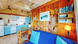 A cozy cabin interior with a wooden wall, a blue sofa, a dining table, and a kitchen area.