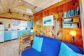 A cozy cabin interior with a wooden wall, a blue sofa, a dining table, and a kitchen area. - Charming Private Truro Cottage with Deck Near Bay Beaches (Truro)