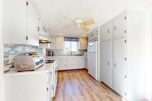 Una cocina con armarios blancos, un ventilador de techo, un tostador y un letrero que dice 'BEACH HOUSE'.