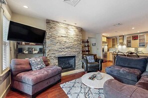 A living room with a stone fireplace, leather sofas, a flat-screen TV, and a kitchen in the background. - Modern west end condo close to beach with deck, W/D, parking, & AC (Provincetown)