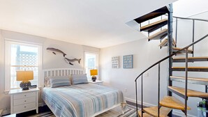 Quarto com cama, mesas de cabeceira, abajur, relógio na parede, um tubarão decorativo, escada e uma frase motivacional na parede.