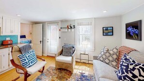 Una sala de estar con un sofá, un sillón y una mesita. Se ven dos ventanas con persianas, una puerta y una zona de cocina.