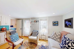 A living room with a sofa, armchair, and a small table. There are two windows with blinds, a door, and a kitchen area visible. - Sunset Serenade | Nautical condo in Provincetown (Provincetown)