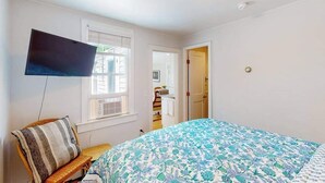 Un dormitorio con una cama, un televisor montado en la pared, una ventana y una puerta que conduce a otra habitación.