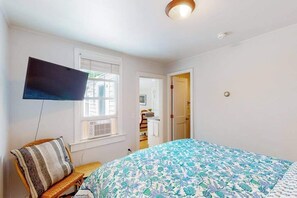 A bedroom with a bed, a TV mounted on the wall, a window, and a door leading to another room. - Sunset Serenade | Nautical condo in Provincetown (Provincetown)
