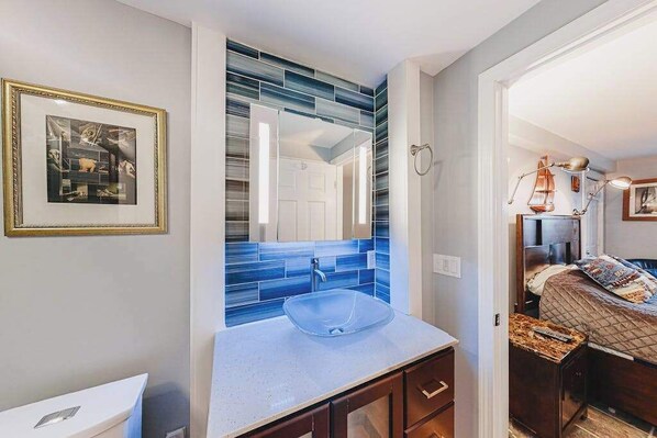 A bathroom with a glass sink, a framed picture on the wall, and a blue-tiled backsplash. - Beautiful condo near the ocean with heated pool, hot tub, grills and kitchenette (Provincetown)