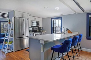 A modern kitchen with stainless steel appliances, a central island with bar stools, and a window with blue trim. - Gorgeously redesigned 1830 home in the heart of town - walk everywhere (Provincetown)