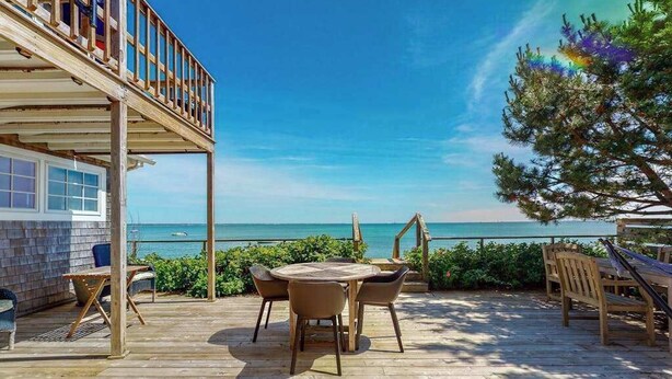 A wooden deck with a round table and chairs, overlooking a clear blue sea.