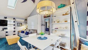Una sala de estar moderna con una mesa blanca, sillas blancas, una lámpara de techo, una estantería y un televisor.