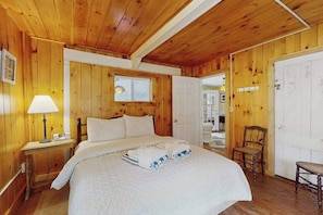 A wooden-paneled room with a bed, a nightstand, a chair, and a door. - Charming home with walkable beach, large yard, deck, fire pit, and A/C (Oak Bluffs)