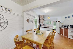 A dining area with a wooden table and chairs, a kitchen in the background, and a compass on the wall. - Charming home with walkable beach, large yard, deck, fire pit, and A/C (Oak Bluffs)