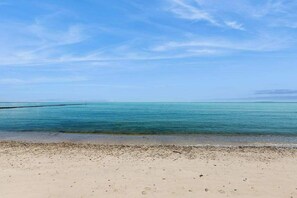 En strand med klart vatten, en sandstrand och en brygga en bit bort.