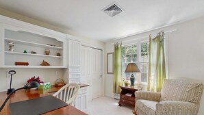 Une pièce comprenant une table à manger en bois, une chaise, une lampe, un canapé, une fenêtre avec des rideaux et un mur blanc avec des étagères.