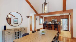 Un coin repas avec une table en bois, des chaises, un miroir rond et une cuisine en arrière-plan.