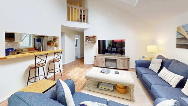 Ein modernes Wohnzimmer mit einer Couch, einem Hocker und einem Fernseher. Es gibt eine Küchenzeile mit einer Arbeitsplatte und Barhocker. Im Hintergrund ist ein Balkon zu sehen.