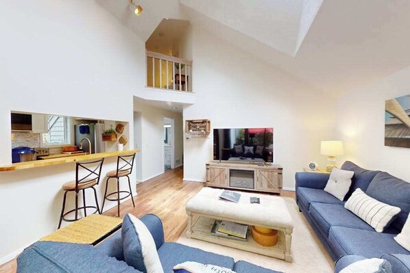 Un salon moderne avec un canapé, un pouf et un téléviseur. On aperçoit un coin cuisine avec un comptoir et des tabourets. Un balcon est visible au fond.
