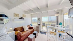 A modern, well-lit room with a brown leather sofa, a dining table with chairs, a TV, and a large window offering a view of the outdoors. - Ocean-view studio near golf with patio, beach gear, kitchenette, & partial AC (North Truro)
