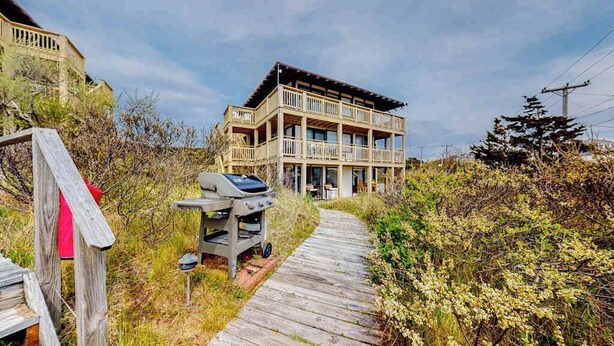 A multi-story building with a wooden deck leading to the entrance, a barbecue grill, and a pathway surrounded by vegetation. - Ocean-view studio near golf with patio, beach gear, kitchenette, & partial AC (North Truro)