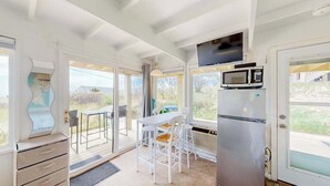 A kitchen with a refrigerator, microwave, and a television. There is a dining table with chairs and a view of the outdoors through large windows. - Ocean-view studio near golf with patio, beach gear, kitchenette, & partial AC (North Truro)