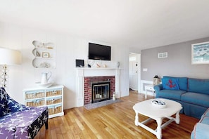 A living room with a fireplace, a television mounted above it, a blue sofa, and a white coffee table. - Renovated, river view home with yard games - walk to town dock & beach (West Dennis)