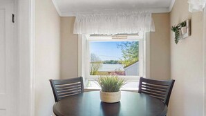 A dining area with a round table, two chairs, a potted plant, a window with a view of greenery, and a white door.