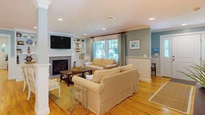 A living room with a fireplace, a television, and a dining area with chairs and a table. - Gorgeously rebuilt Cape Cod estate with lovely grounds, walk to private beach (West Yarmouth)