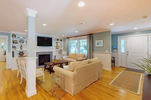 A living room with a fireplace, a television, and a dining area with chairs and a table. - Gorgeously rebuilt Cape Cod estate with lovely grounds, walk to private beach (West Yarmouth)