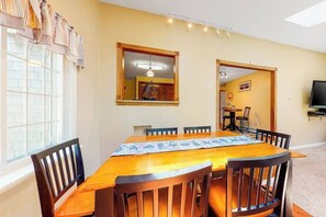 A dining area with a wooden table and chairs, a window with curtains, a mirror with a ship decoration, and a television on the wall.