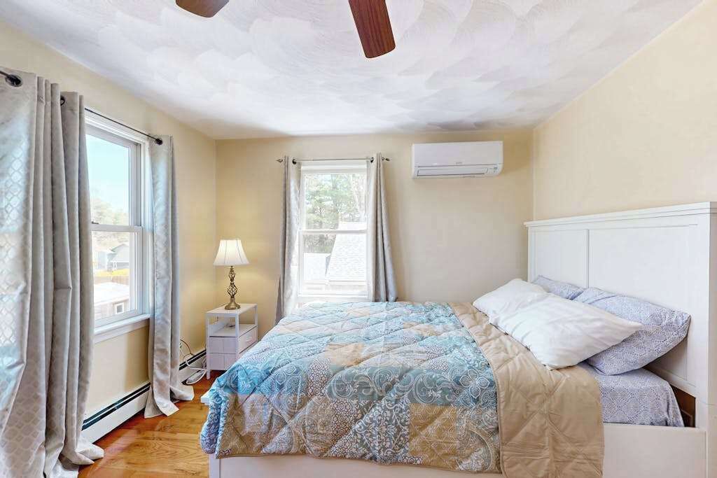 Une chambre à coucher comprenant un lit, une table de chevet, un appareil de climatisation et deux fenêtres avec des rideaux.