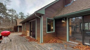 A wooden deck with a barbecue grill, a house with a green trim, and a glass door. - Gorgeous home near the ocean w/ a full kitchen, deck, & large, fenced back yard (Vineyard Haven)