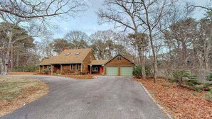 A two-story house with a garage, surrounded by trees and a driveway. - Gorgeous home near the ocean w/ a full kitchen, deck, & large, fenced back yard (Vineyard Haven)