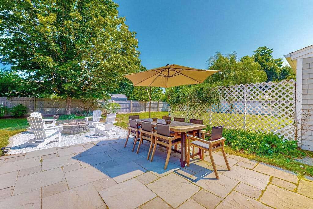 Eine Terrasse im Freien mit einem Holztisch und Stühlen, einem weißen Gitterzaun und einem großen Baum.