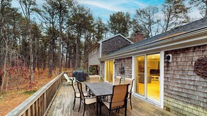Eine Terrasse mit Tisch und Stühlen, einem überdachten Grill und einem Haus mit Holzfassade und Glasüren.