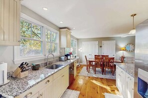 A kitchen with granite countertops, wooden cabinets, a dining area with chairs, and a large window with a view of trees.