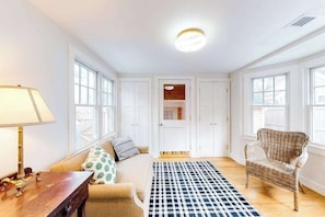 A room with a sofa, a chair, a lamp, a checkered rug, and a door leading to another room. - Cottage w/ water views from front yard, central ac & fireplace (Vineyard Haven)