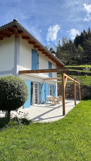 A two-story house with a covered patio, a stone wall, and a view of a hillside.