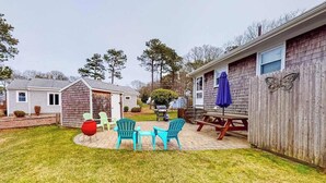 A backyard with a picnic table, chairs, and a grill area. - Cozy cottage, WiFi, A/C, Gas grill, W/D, Firepit (South Yarmouth)
