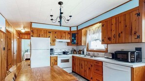 A compact kitchen with wooden cabinets, a dining table, a refrigerator, a stove, and a microwave. - Cozy cottage, WiFi, A/C, Gas grill, W/D, Firepit (South Yarmouth)
