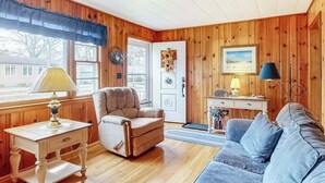 A cozy room with wooden walls, a beige armchair, a blue sofa, a small desk with a lamp, and a door with a wreath. - Cozy cottage, WiFi, A/C, Gas grill, W/D, Firepit (South Yarmouth)