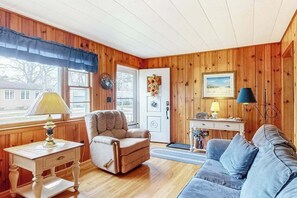 A cozy room with wooden walls, a beige armchair, a blue sofa, a small desk with a lamp, and a door with a wreath. - Cozy cottage, WiFi, A/C, Gas grill, W/D, Firepit (South Yarmouth)